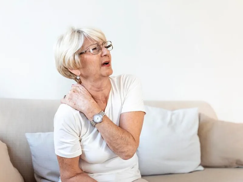 An older woman sitting on a sofa holding her shoulder in discomfort